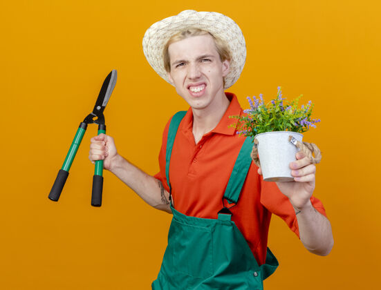 剪子失望的年轻园丁穿着连体衣 戴着帽子 拿着树篱剪和盆栽植物 站在橙色的背景上 看着摄像机不高兴篱笆立场相机