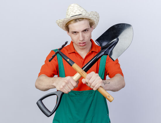 铲子年轻的园丁 穿着连体衣 戴着帽子 拿着小耙子和铲子 严肃地双手交叉站在白色背景上看着摄像机看连身衣站着