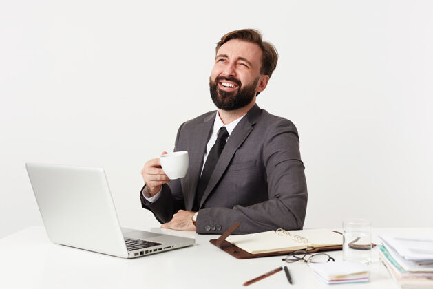 领带一张室内照片 一个快乐的年轻的留着胡子的黑发男人拿着他的笔记本电脑在办公室工作 一边喝茶一边开心地笑着 穿着灰色的西装 打着白色的领带杯子积极正式