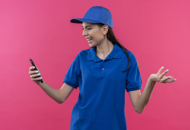 粉色穿着蓝色制服 戴着帽子的年轻送货女孩看着智能手机的屏幕失望了制服送货看起来