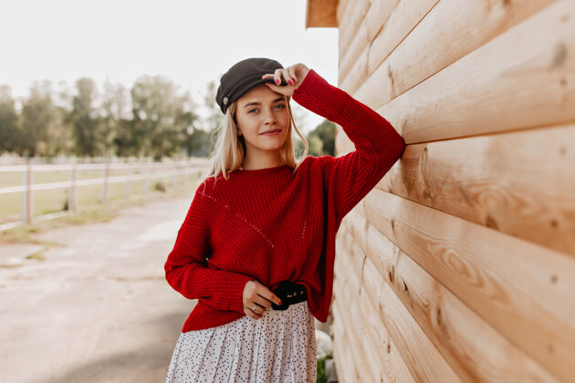 木头房子迷人的金发碧眼 自然的妆容 温柔的表情穿着红色套头衫 戴着时髦帽子的年轻女子在木屋附近摆姿势漂亮身体魅力