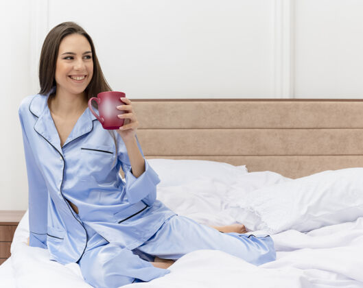 蓝色穿着蓝色睡衣的年轻漂亮女人坐在床上 端着一杯咖啡 面带微笑 坐在卧室的室内灯光背景下咖啡睡衣漂亮