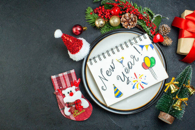 圆锥体上图为餐桌上带笔的螺旋笔记本圣诞树杉木树枝针叶树圆锥形礼品盒圣诞老人帽圣诞袜在黑色背景的左侧左边壁板视图