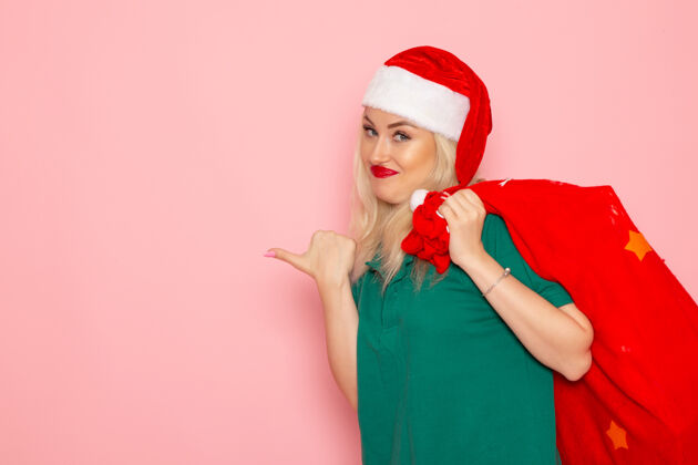 肖像正面图年轻女性拎着红包在粉色墙上赠送节日模特新年彩照圣诞老人美丽圣诞老人微笑