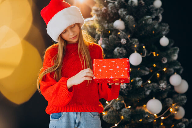 圣诞树可爱的少女戴着圣诞树旁的红色圣诞帽爱时尚红色