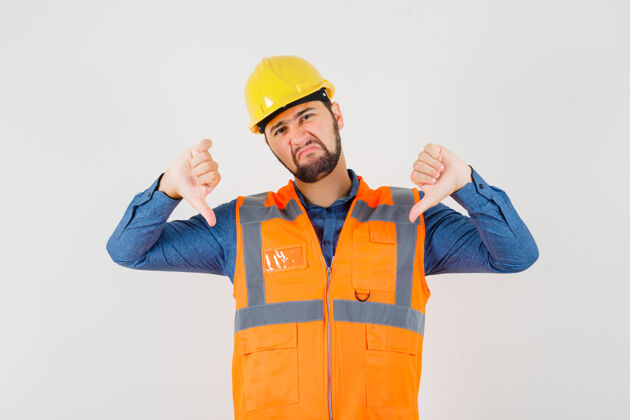 工人年轻的建筑工人在衬衫 背心 头盔上竖起大拇指 看上去很不满意男性肖像工匠