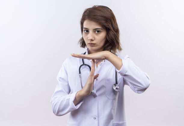 医生穿着白大褂的年轻女医生戴着听诊器 看上去很累 用手做着暂停的手势时间看外套