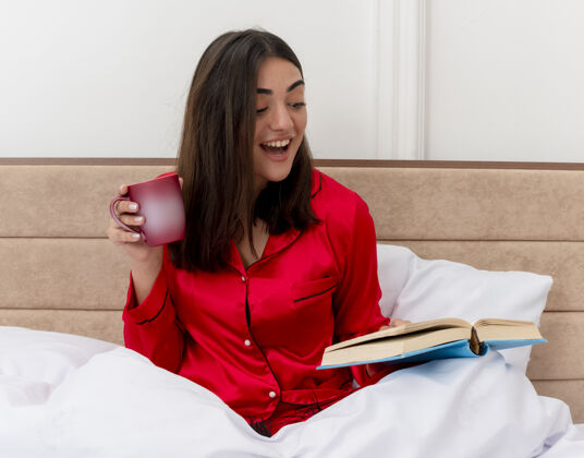 放松穿着红色睡衣的年轻漂亮女人坐在床上 喝着咖啡 看书 放松地享受周末 在卧室的室内灯光背景下 面带笑容地享受着快乐快乐咖啡睡衣