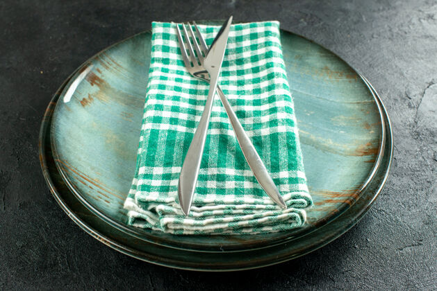 盾牌底视图交叉餐刀和餐叉在绿色和白色方格餐巾在盘子上黑色的桌子上盘子十字架碗