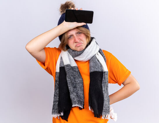 温暖不健康的年轻女子穿着橙色t恤 脖子上围着暖和的围巾 戴着帽子 感觉很糟糕 拿着智能手机和纸巾 患感冒和流感 在白墙上看起来很失望移动流感抱着