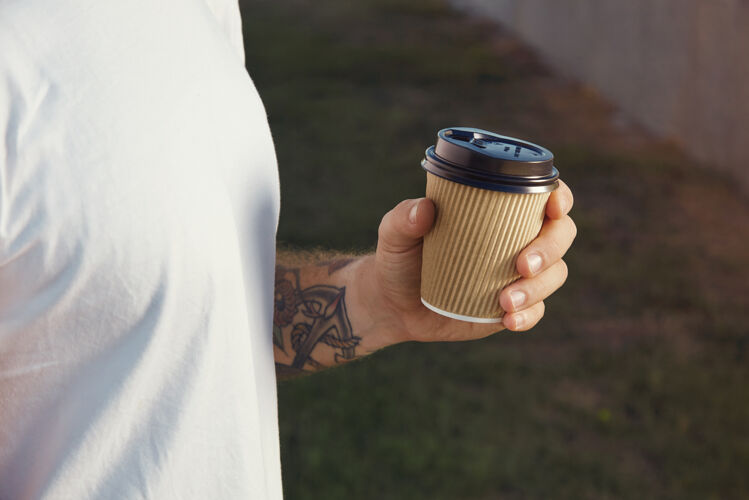 创意手和胸部的一个白色纹身男子身穿白色无标签t恤持有浅棕色纸咖啡杯茶杯子城市