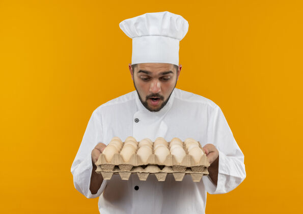 纸箱惊讶的年轻男厨师穿着厨师制服 拿着一盒鸡蛋看着孤立在橙色空间里鸡蛋举行男性