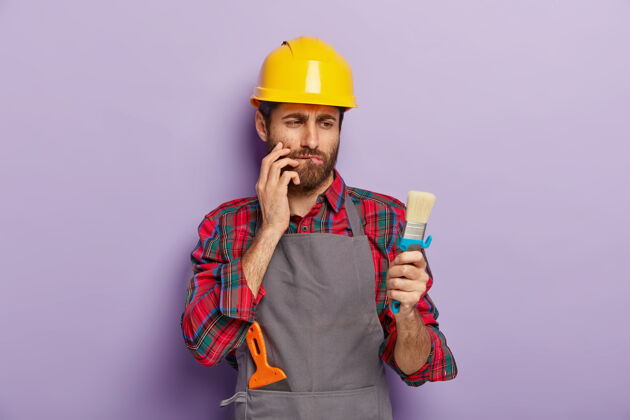 室内墙画概念认真周到的工匠看着干净的刷子 咬着下唇 想着如何开始他的公寓装修 戴着黄色的头饰和围裙 孤立在紫色的墙上人修理工作