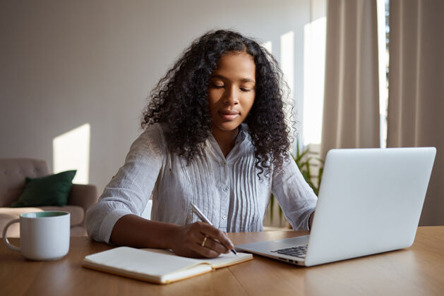 笔记本室内照片：一位年轻漂亮的黑皮肤女性卷发 坐在办公桌前 对着打开的便携式电脑 一边喝着咖啡 一边在抄写本上写下一天的计划微笑笔记本电脑幸福