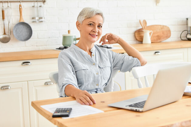 老年人现代精力充沛的成年女商人坐在餐桌旁吃早餐 用便携式电脑查看电子邮件时尚的高级女性自由职业者在家里用笔记本电脑工作老年人无线个人电脑