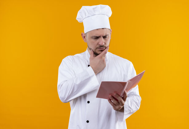 垫着体贴的年轻帅哥厨师穿着厨师制服 手放在下巴上 手拿着笔记本看着橙色的空间制服笔记年轻