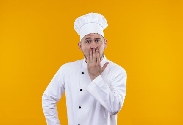 厨师惊讶的年轻帅哥厨师穿着厨师制服把手放在嘴上孤立的橙色空间放嘴手