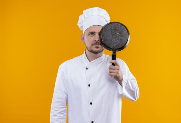 年轻年轻帅气的厨师穿着厨师制服 手里拿着煎锅 躲在它后面 看起来孤立在橙色的空间里拿着厨师制服