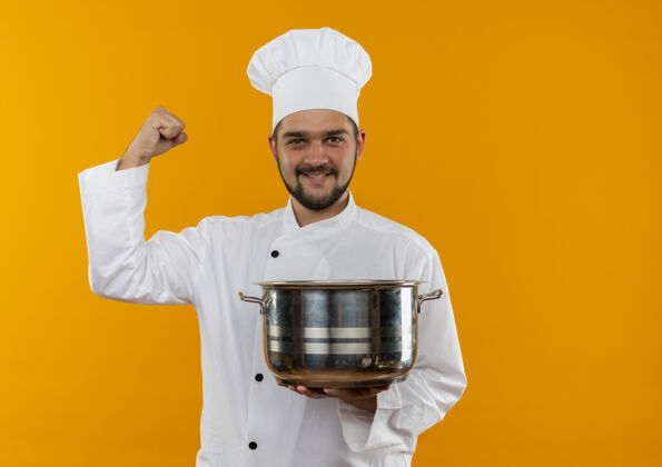 男性身着厨师制服的年轻男厨师微笑着拿着锅 在橙色的空间里做着强烈的孤立的手势强壮手势微笑