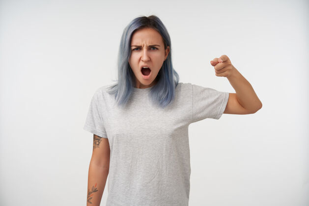 蓝发心怀不满的年轻蓝发纹身女性 穿着灰色基本款t恤 站在白色地面上愤怒地竖起食指女人室内肖像
