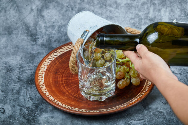 串小桶葡萄放在陶瓷盘子里 用手把葡萄酒倒进大理石上的玻璃杯里桌子深色绿色