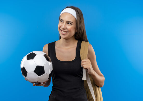 年轻面带微笑的年轻漂亮的运动女孩戴着头带和腕带和背包拿着足球 看着蓝色空间上孤立的一面头带复制蓝色