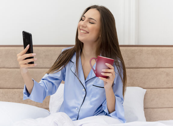 坐着穿着蓝色睡衣的年轻美女坐在床上喝着一杯咖啡 用智能手机微笑着 在卧室内部的灯光背景下露出幸福的脸睡衣女人智能手机