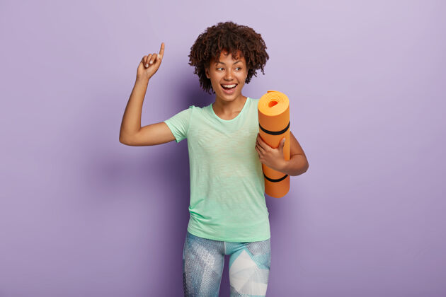 锻炼快乐微笑无忧无虑的卷发非裔美国女运动员携带皱巴巴的瑜伽垫 抬起手臂并向上表示 享受良好的训练 穿着t恤和紧身裤运动概念休闲人身材
