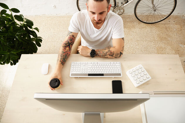 开始顶视图纹身的自由职业者拿着咖啡从外卖纸杯 而在他的电脑显示器上看与midi音乐控制器在家庭桌面上智能手机上在线家女人