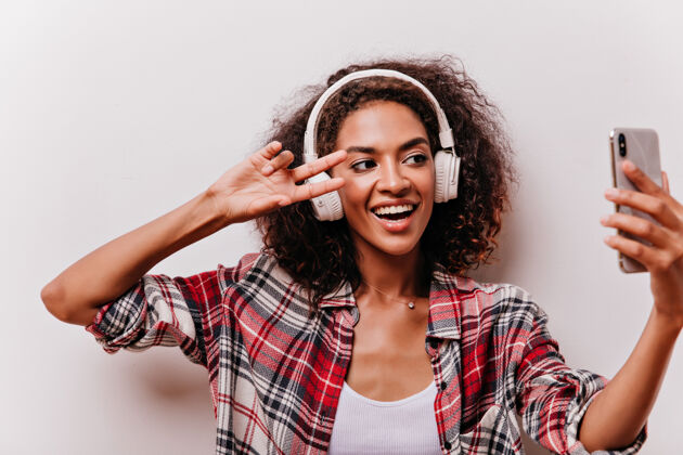成人黑眼积极的女人自拍和平标志和笑快乐的卷发女孩在大耳机玩得开心Mp3耳机表情