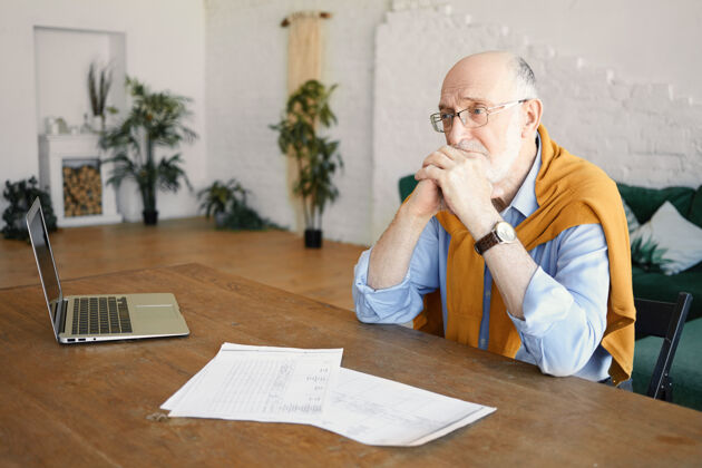 财务在室内拍摄的照片中 一位愁眉苦脸的大胡子男性企业家坐在办公桌旁 手里拿着笔记本电脑和报纸 表情沮丧 对财务问题感到沮丧 双手放在下巴下面不快乐思考悲伤