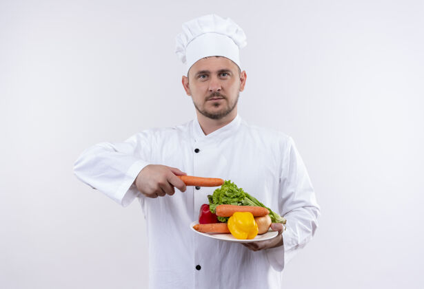 蔬菜年轻帅气的厨师身着厨师制服 端着盛有蔬菜的盘子 用胡萝卜指着他们 看着孤立的白色空间制服指着胡萝卜