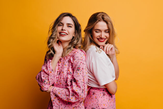 享受穿着粉色上衣的梦幻女孩和她的妹妹合影可爱的女性朋友在黄色的墙上笑着朋友表情微笑