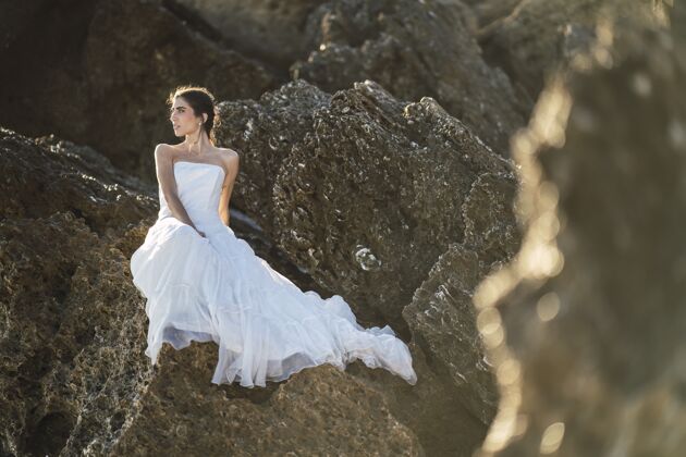 地平线选择性聚焦拍摄一位身穿白色连衣裙的黑发女性在岩石上摆姿势日落深色海洋