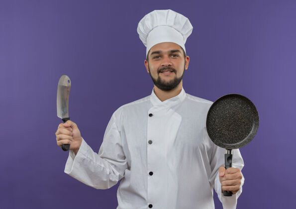 男身着厨师制服 面带微笑的年轻男厨师手拿菜刀和煎锅孤立在紫色的空间里平底锅切肉刀微笑