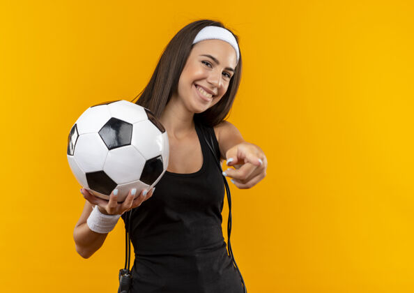 球微笑着的年轻漂亮的运动女孩戴着头带和腕带拿着足球 用跳绳指着脖子孤立在橙色的空间女孩橙色指向