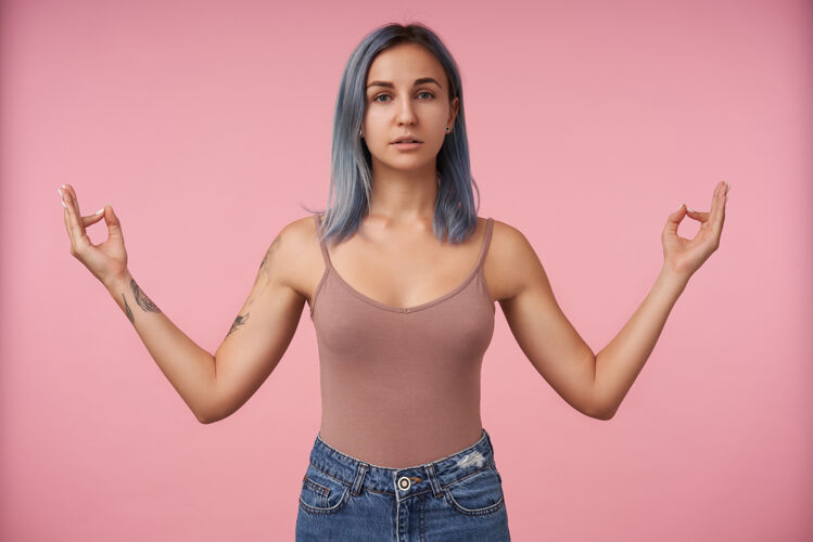 心情年轻女性的肖像 蓝色短发卷曲 手指高举 站在粉色米色衬衫上的纳马斯特标志发型灰色冥想
