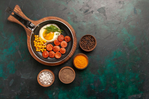 早餐顶视图深绿色背景上的炒鸡蛋 香肠和不同的调味品调味品健康蔬菜