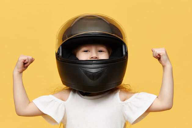 摩托车手一张小女孩赛车手戴着黑色安全摩托车头盔对着黄色墙壁摆姿势的照片 展示了她的二头肌人 极限运动和肾上腺素的概念自行车保护设备