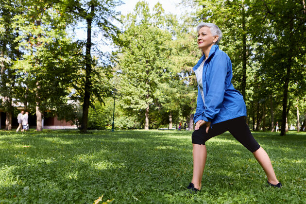微笑精力充沛的女性退休人员穿着时尚的运动服 选择健康积极的生活方式 在森林或公园的绿草上训练 做弓箭步 拥有快乐快乐的容颜老年人 健康和夏天草地老年人森林