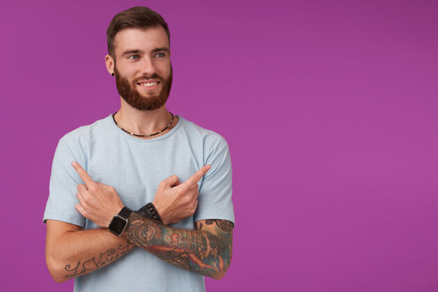 纹身长相讨人喜欢的年轻纹身男性 胡须欢快地笑着 食指指向不同的侧面 穿着休闲服站在紫色的地面上蓝色手表手