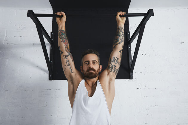 活跃残忍的纹身运动男子在白色无标签坦克t恤显示健美动作的经典拉起近距离挂在拉杆上 看着相机健康强壮肌肉