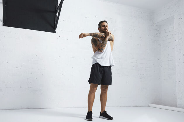 健康运动纹身男子在白色空白坦克t恤伸展他的肱二头肌在手臂上锻炼后 在砖墙旁边的黑色拉杆隔离健身房动作交叉