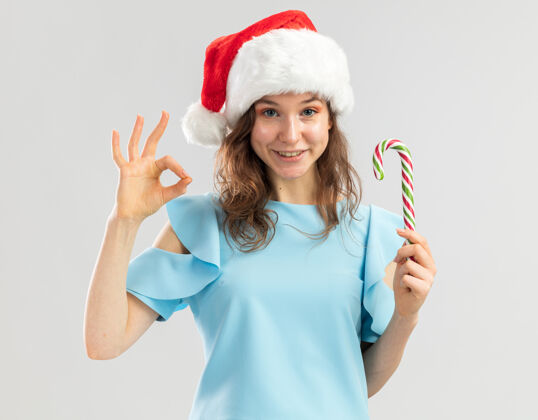 积极穿着蓝色上衣 戴着圣诞帽 手持糖果手杖的年轻女子看起来很开心 积极地表示同意帽子举行站
