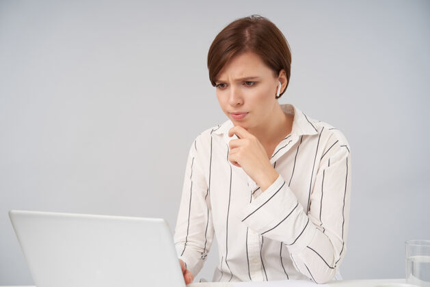 模特困惑的棕色头发的年轻女士 自然化妆 抬起手托着下巴 皱着眉头 沉思地看着她的笔记本电脑屏幕 孤立在白色屏幕上短衣服耳机