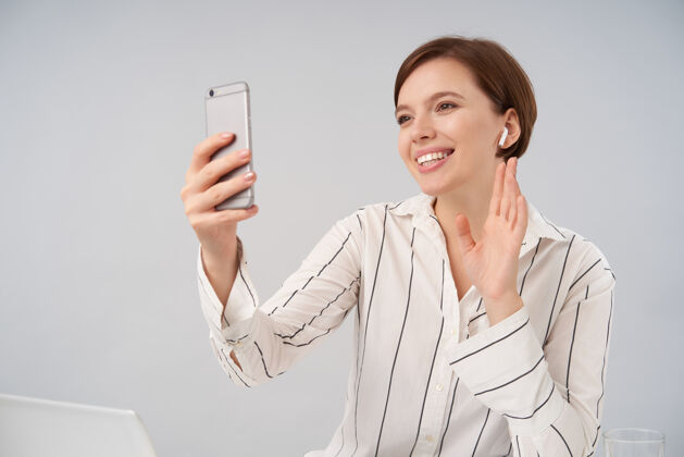休闲快乐的年轻短发黑发女子 自然妆容 举手打招呼 面带微笑 用智能手机进行视频通话 与白色隔离女性电话笔记本电脑