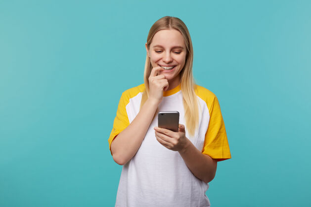 牙齿一幅年轻的金发长发女人的肖像 她穿着休闲服 在智能手机屏幕上若有所思地看着 脸上带着积极的微笑 与蓝色隔绝基本发型电话