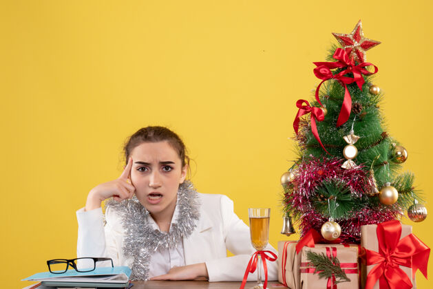 女医生正面图：女医生坐在黄色背景的桌子后面 还有圣诞树和礼品盒插花新郎花