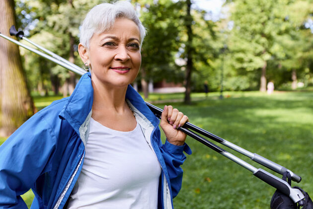 老年人一位时尚自信的欧洲女士 灰色短发 肩上扛着北欧手杖 站在松林中 做完有氧运动回家 面带微笑徒步旅行活动老年人