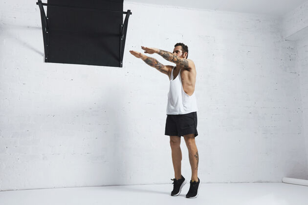 体操强壮的纹身在白色无标签坦克t恤男运动员显示健美操动作蹲小腿上升 顶端位置健身房运动拉